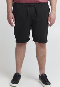 Homme portant un short noir retroussé avec une taille à cordon et des baskets noires, debout devant un fond clair uni.