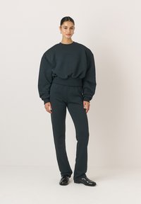 Ciemnozielona bluza z długim rękawem i dopasowane spodnie typu tapered, wykonane z miękkiego materiału. Posiada okrągły dekolt i ściągacze przy rękawach, noszone z czarnymi butami.