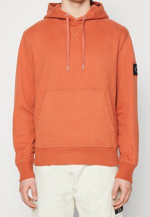 Hombre vistiendo una sudadera con capucha naranja con bolsillo frontal y cordones, combinada con pantalones cargo blanco roto.