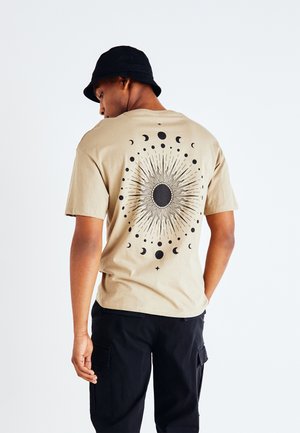 Beige cotton t-shirt with a black celestial graphic featuring sun, moons, and stars. Short sleeves, relaxed fit, and crew neckline.