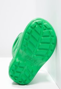 Chaussure en caoutchouc verte Crocs vue par le dessous, présentant une semelle texturée avec de grands motifs de relief et des détails de logo.