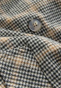 Primer plano de una tela de patrón houndstooth en tonos beige, negro y marrón, con un gran botón marrón de cuatro agujeros y detalle de bolsillo cosido.
