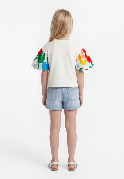 Jauna meitene ar gariem blondiem matiem stāv ar muguru pret skatītāju, valkājot baltu kreklu ar krāsainiem ziedu pumpainiem piedurkņiem, gaišas džinsa šortus un baltas sandales.