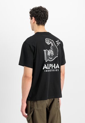 Uomo con capelli ricci che indossa una t-shirt nera con grafica bianca di un braccio che si flette e la scritta "ALPHA INDUSTRIES" sulla schiena, abbinata a pantaloni marroni.