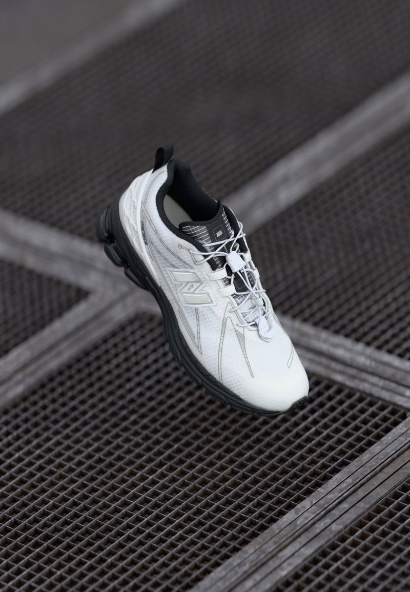 Chaussure de sport blanche et noire avec lacets élastiques sur une surface en grille métallique sombre avec des lignes croisées en arrière-plan.