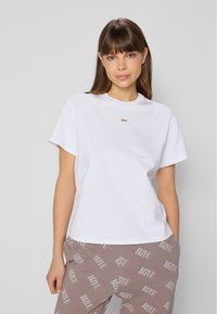 Biała koszulka z krótkim rękawem i okrągłym dekoltem, z małym złotym logo z przodu, w zestawie z brązowymi z wzorem spodniami dresowymi.