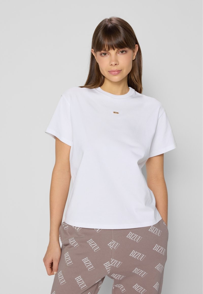 Biała koszulka z krótkim rękawem i okrągłym dekoltem, z małym złotym logo z przodu, w zestawie z brązowymi z wzorem spodniami dresowymi.