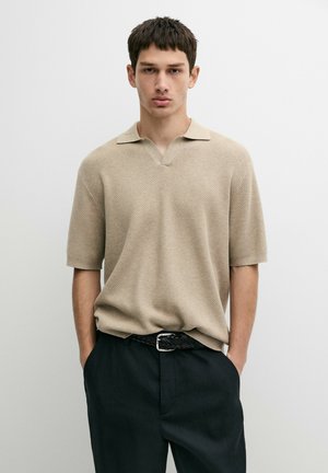 Junger Mann steht mit den Händen in den Taschen, trägt ein beiges, strukturiertes Kurzarmhemd und schwarze Hose vor einem schlichten hellen Hintergrund.
