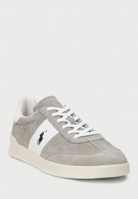 Grå mockaskor med vita detaljer och snörning. Har en logotyp och texturerad detaljering på sidan. Gummisula, klassisk sportig design.