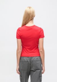 Γυναίκα με μακριά ξανθά μαλλιά, γυρισμένη πίσω, που φορεί εφαρμοστό κόκκινο κοντομάνικο πουκάμισο και χαλαρό γκρι παντελόνι καρό με πίσω τσέπες.