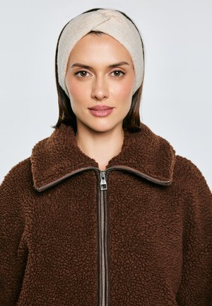 Kobieta w jasnobeżowej dzianinowej opasce na głowę i brązowej polarowej kurtce zapinanej na zamek na tle jasnego, jednolitego tła.