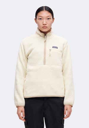Mladá osoba s rovnými tmavými vlasmi nosí krémový fleece pulóver so zipsom a logom na hrudi, v kombinácii s čiernymi nohavicami.