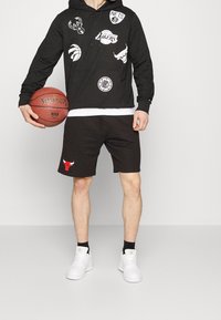 Henkilö pitää käsissään koripalloa, pukeutuneena mustaan huppariin, jossa on NBA-joukkueiden logoja, mustiin shortseihin, joissa on Chicago Bullsin logo, valkoisiin tennareihin ja mustiin sukkiin.