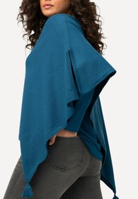 Poncho teal realizzato in tessuto morbido, con una vestibilità ampia, taglio diagonale e pon pon decorativi agli angoli. Abbinato a jeans grigio scuro.