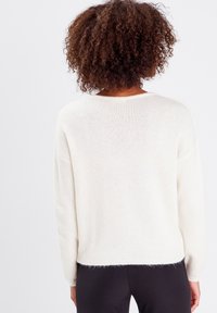Pull en tricot blanc avec une coupe décontractée, des manches longues et un ourlet côtelé. Le tissu a une texture douce et une finition légèrement duveteuse.