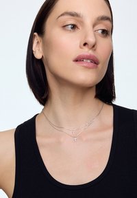 Srebrne, warstwowe naszyjniki z małymi wisiorkami w kształcie krzyża. Posiadają delikatne łańcuszki i błyszczące wykończenie, noszone na czarnej bluzce na ramiączkach.