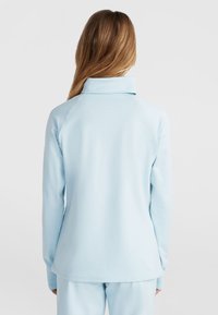 Kobieta z długimi blond włosami, ubrana w jasnoniebieską bluzkę z długim rękawem i wysokim kołnierzem, pokazana od tyłu na jednolitym tle.