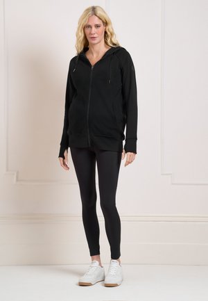 Lyshåret kvinde iført en sort hættetrøje med lynlås, sorte leggings og hvide sneakers, der står mod en ensfarvet beige væg.