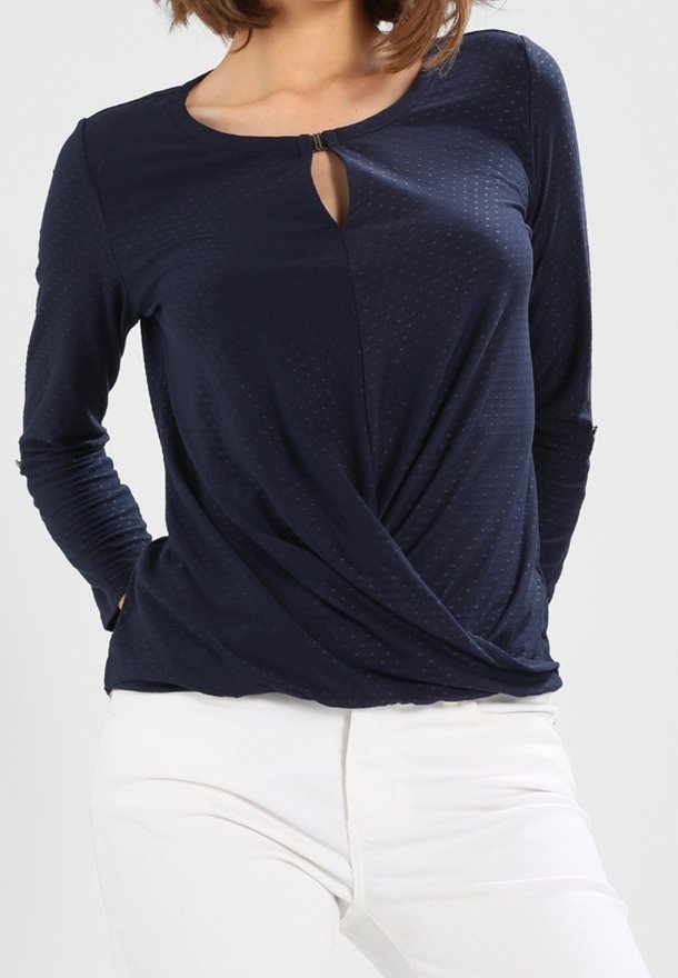 Nainen, jolla on yllään tummansininen pitkähiha toppi, jossa on pieni avainaukko edessä ja kiertävä helma, yhdistettynä valkoisiin housuihin.