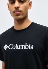 Jeune homme portant un t-shirt noir à col rond avec le logo et l'emblème blancs "Columbia" sur la poitrine, sur un fond uni.