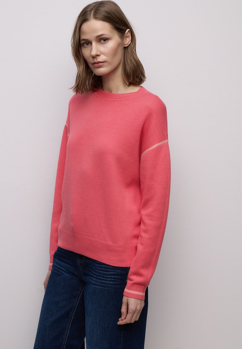Femme aux cheveux bruns mi-longs portant un pull corail rose et un jean bleu foncé, debout devant un mur gris clair uni.