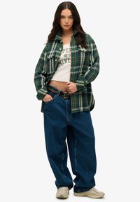 Chemise en flanelle à carreaux verts et bleus, haut court blanc, et jeans bleus amples. Accentué par une ceinture noire cloutée et des baskets neutres.