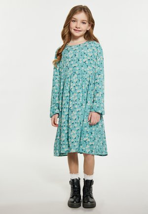 Langärmliges türkisfarbenes Kleid mit floralem Muster, gerafftem Bund und ausgestelltem Rock. Kombiniert mit schwarzen Schnürstiefeln und weißen Rüschenstrümpfen.