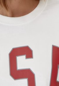 Camiseta de algodón blanca con gran texto rojo "USA" contorneado en gris. Cuello redondeado y textura suave visible en el primer plano de la tela.