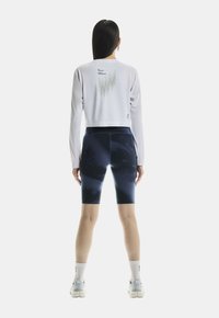 Donna in piedi di spalle, con un crop top bianco a maniche lunghe con testo e pantaloni da ciclismo navy tie-dye, calze bianche e scarpe da ginnastica bianche.