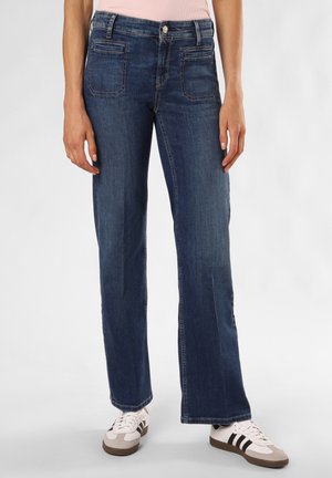 Jeans en denim bleu à jambe large avec poches avant et arrière, passants de ceinture, couture visible et léger délavage, associés à des baskets blanches et marron.