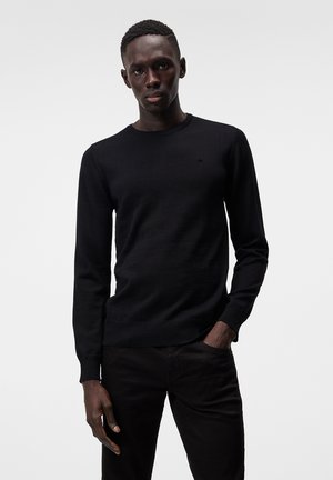 Schwarzer Strickpullover mit rundem Halsausschnitt, gerippten Bündchen und Saum, kombiniert mit dunklen Hosen. Minimalistisches Logodesign auf der Brust.