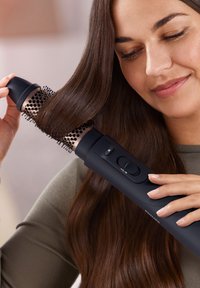 Outil de coiffure avec un embout brosse cylindrique, corps noir, poils texturés et bouton de commande. Les cheveux coiffés sont lisses et brillants.