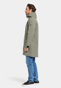 Veste imperméable longue de couleur vert olive avec capuche, manches ajustables et un patch logo noir. Portée avec un jean bleu et des baskets noires.