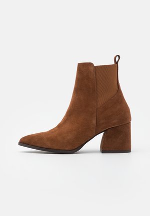 Bottine en daim marron avec un bout pointu et un talon épais. Elle comporte des panneaux latéraux élastiques et une languette arrière pour un enfilage facile.