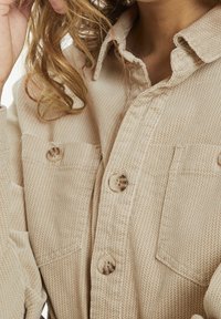 Camicia bottone color beige con motivo, realizzata in tessuto testurizzato, dotata di due tasche sul petto e bottoni rotondi; colletto rifinito in modo curato.