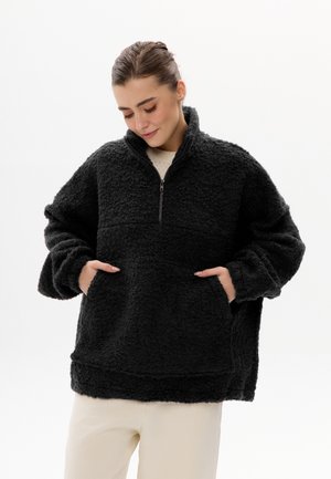 Femme portant un pull polaire texturé noir avec une demi-fermeture éclair et les mains dans les poches avant, associé à un pantalon beige clair.
