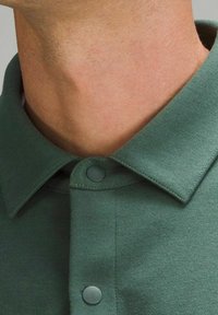 Tejido de polo de color verde oscuro con una textura suave, que presenta un cuello y dos botones grises. Primer plano del cuello y área superior de la prenda.