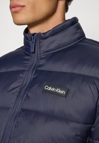 Marineblå puffjakke med høy krage, quiltet design og hel glidelås. Har et svart logo-merke med hvit tekst foran.