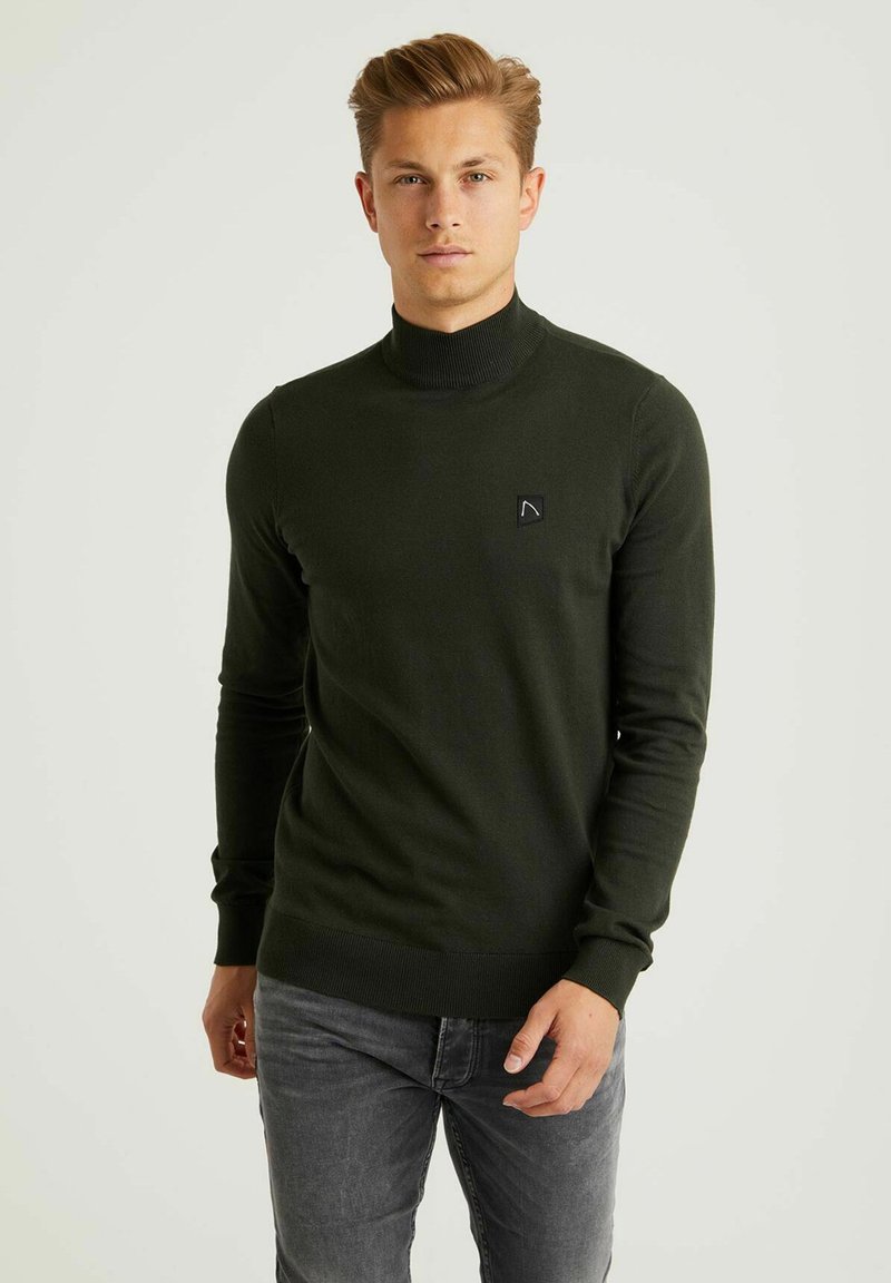 Suéter de cuello alto en verde oscuro, fabricado con un material de punto suave, que presenta un pequeño parche de logo negro en el pecho y puños y dobladillo acanalados.
