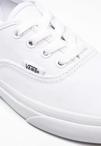 Białe sneakersy Vans z białymi sznurowadłami, metalowymi oczkami i widoczną metką z logo Vans na górnej części z płótna.