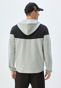 Hellgrauer Kapuzenpullover mit schwarzen Schultereinsätzen. Hergestellt aus weichem Stoff, mit einer Kängurutasche und einer strukturierten Kapuze.