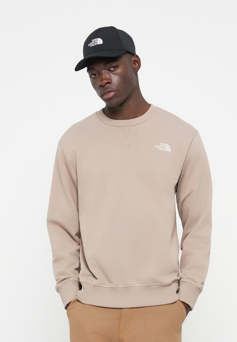 Beige Sweatshirt mit gerippten Bündchen und Halskragen, mit einem weißen Logo. Die schwarze Kappe mit Logo bildet einen Kontrast zu dem hellen Kleidungsstück.