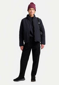 Zwarte gewatteerde jas met een hoge kraag, gedragen over een marineblauw shirt. Zwarte cargobroek en donkere sneakers. Gebreide bordeauxrode muts. Kleine zwarte tas.