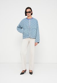 Ανοιχτό μπλε μπουφάν με ραμμένο μοτίβο με διαμάντια, στρογγυλή λαιμόκοψη, τσέπες μπροστά και μαύρα μεταλλικά στοιχεία, συνδυασμένο με κρεμ παντελόνι και μαύρα τακούνια.