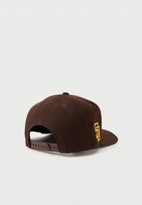 Brown snapback cap with a structured design. Features a yellow "SD" embroidered logo on the side and a plastic adjustable strap.