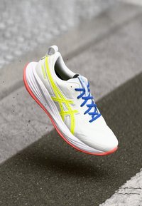 Baskets de course blanches avec des lacets bleus, des accents jaunes et une semelle rose. Présentent un dessus texturé et un design léger.