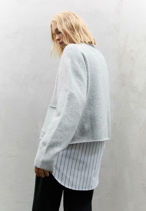 Svijetlosiva skraćena sweatshirt napravljena od mekog materijala, s rebrastim manžetama i opuštenim krojem, preko bijele košulje s vertikalnim prugama.