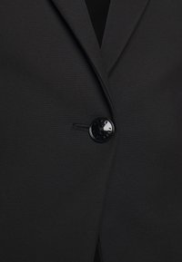 Blazer noir avec une texture lisse, revers crantés et un seul bouton rond noir. Comprend une poche latérale discrète.