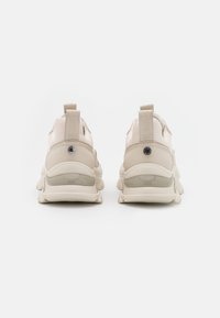 Béžové sportovní tenisky s síťovinovými panely, polstrovaným límcem a robustní gumovou podrážkou. Mají texturovanou patu a logo akcenty.