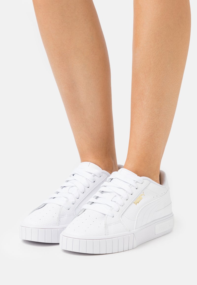 Fehér bőr sneaker, perforált részletekkel, lapos gumitalppal és fehér fűzővel; oldalt diszkrét arany logóval rendelkezik.